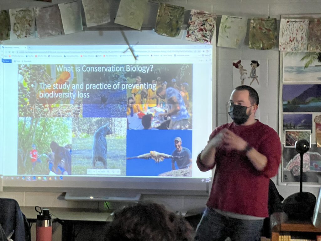 Austin teaching what Conservation Biology is. Picture of Austin in front of a slide that has a bear, humans, and birds.